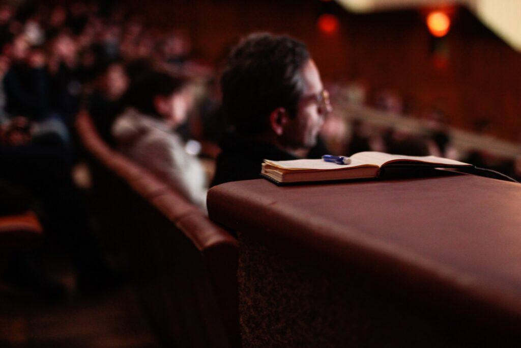 An open notebook with a pen on a brown seat back, with a blurred person and audience in the background.