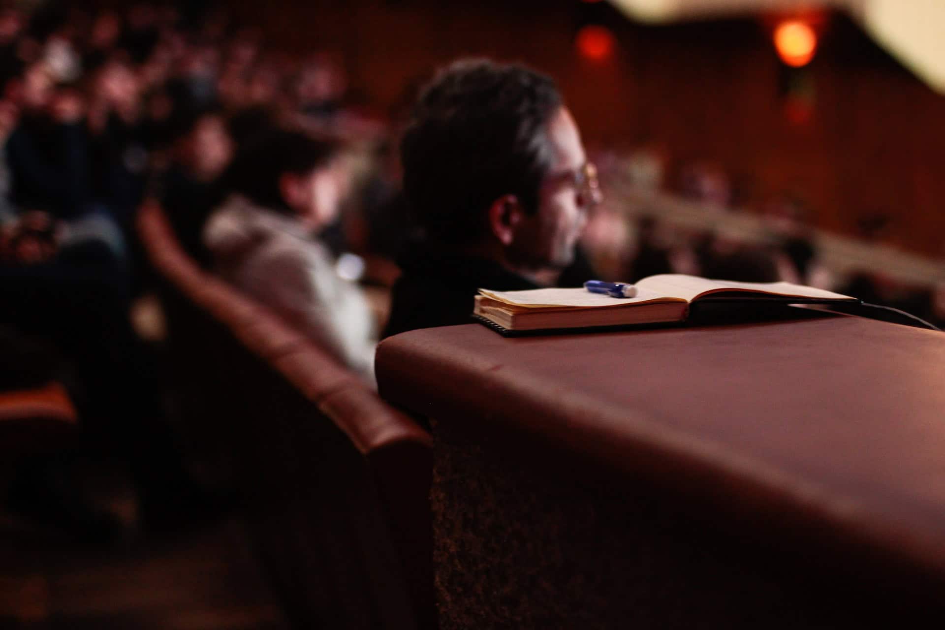 An open notebook with a pen on a brown seat back, with a blurred person and audience in the background.
