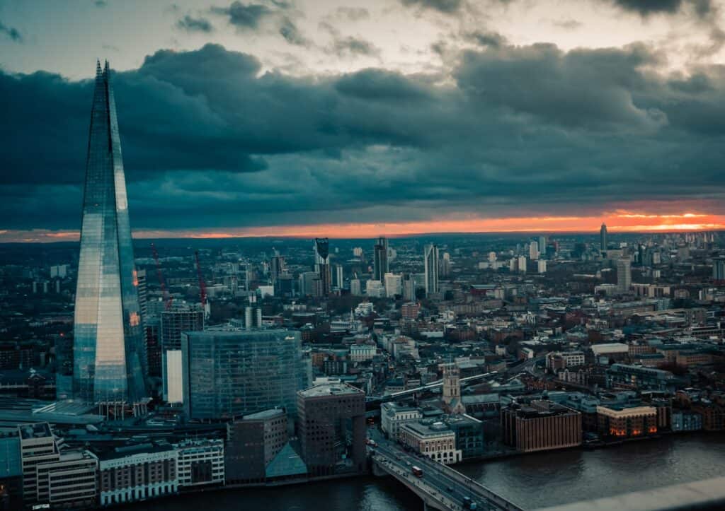 An aerial view of London at dusk, featuring The Shard skyscraper, a river, and a city skyline under a dark, cloudy sky with a vibrant orange sunset.