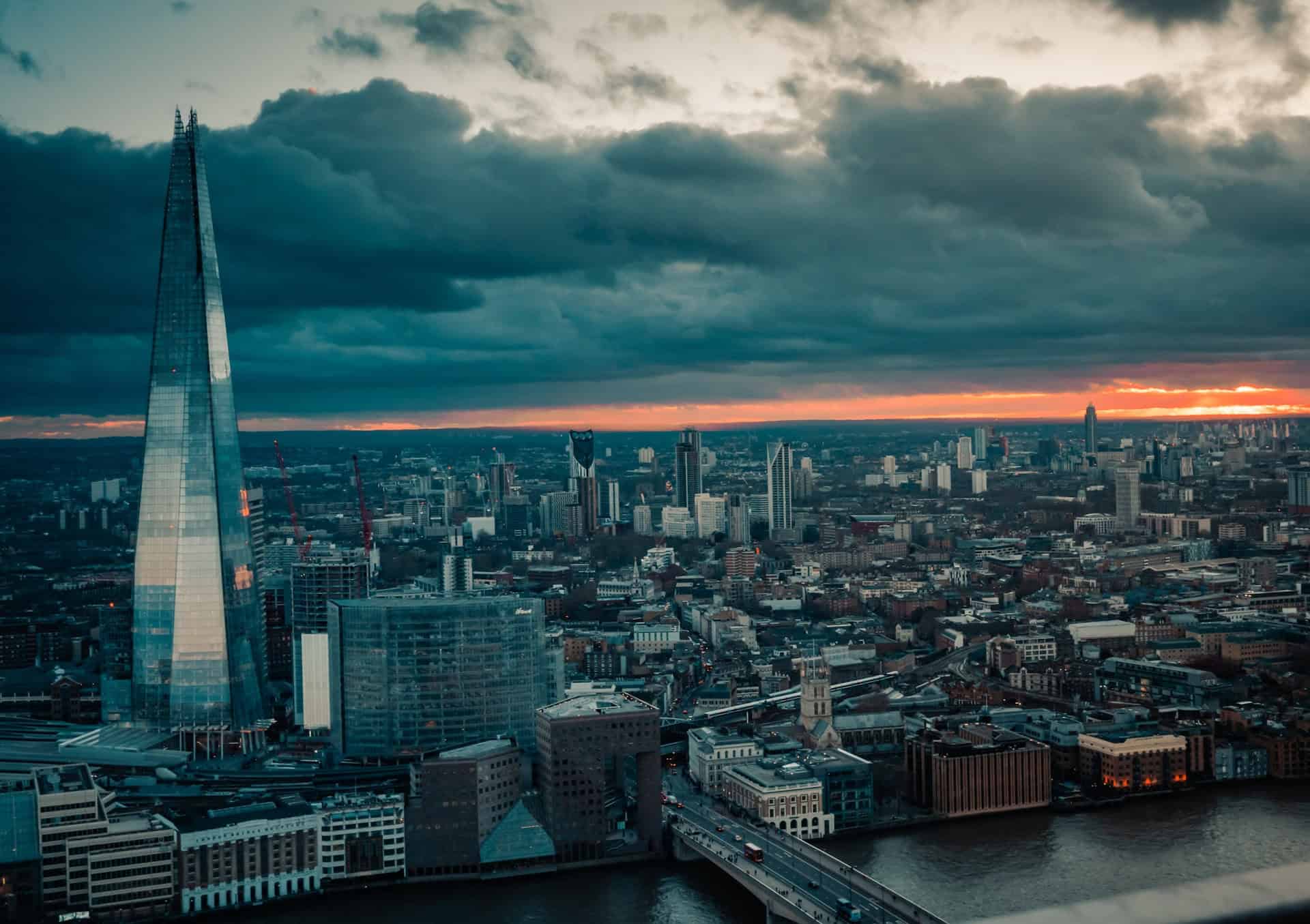 An aerial view of London at dusk, featuring The Shard skyscraper, a river, and a city skyline under a dark, cloudy sky with a vibrant orange sunset.