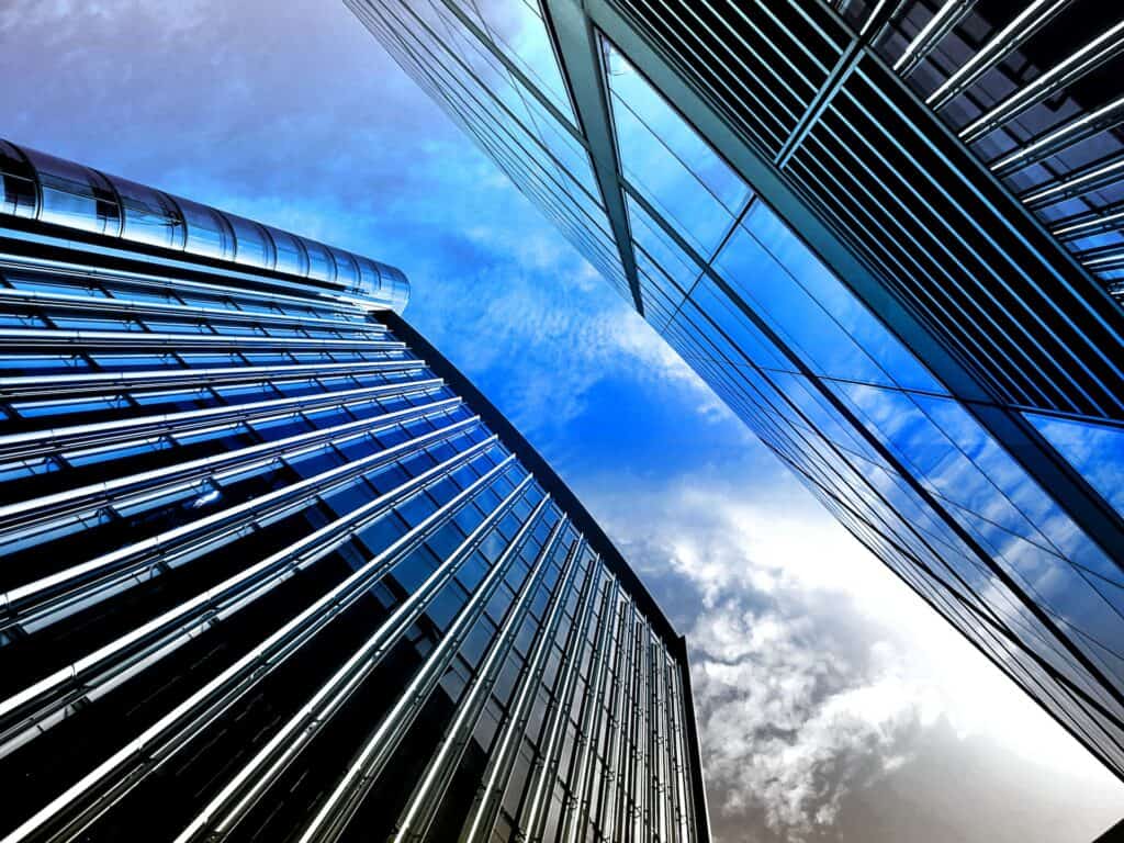 Low-angle view of two modern glass buildings reflecting a blue sky with white clouds.