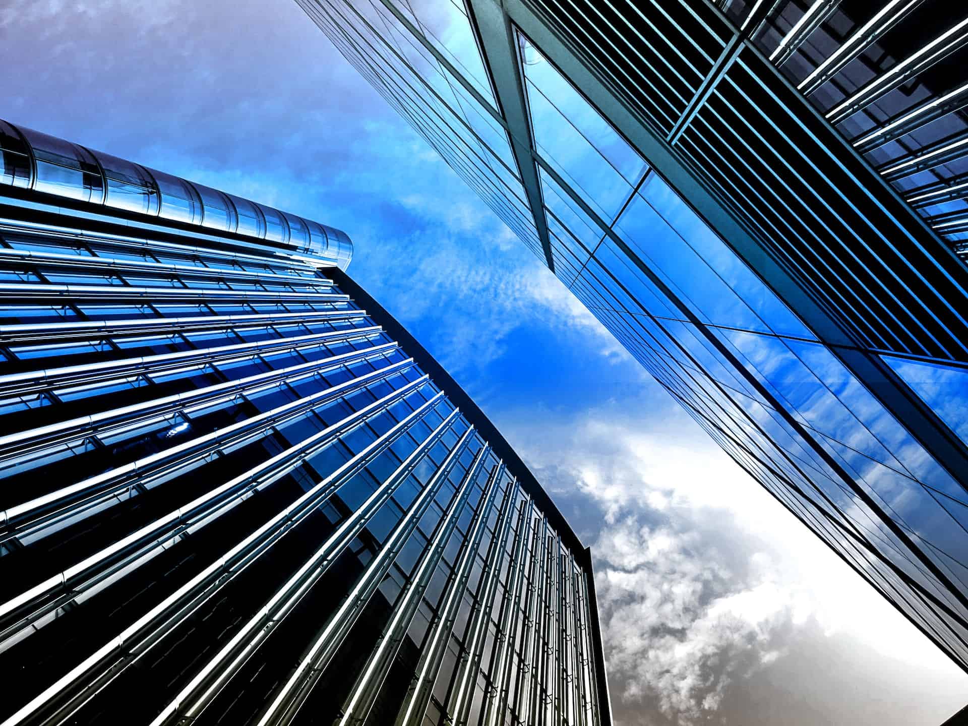 Low-angle view of two modern glass buildings reflecting a blue sky with white clouds.