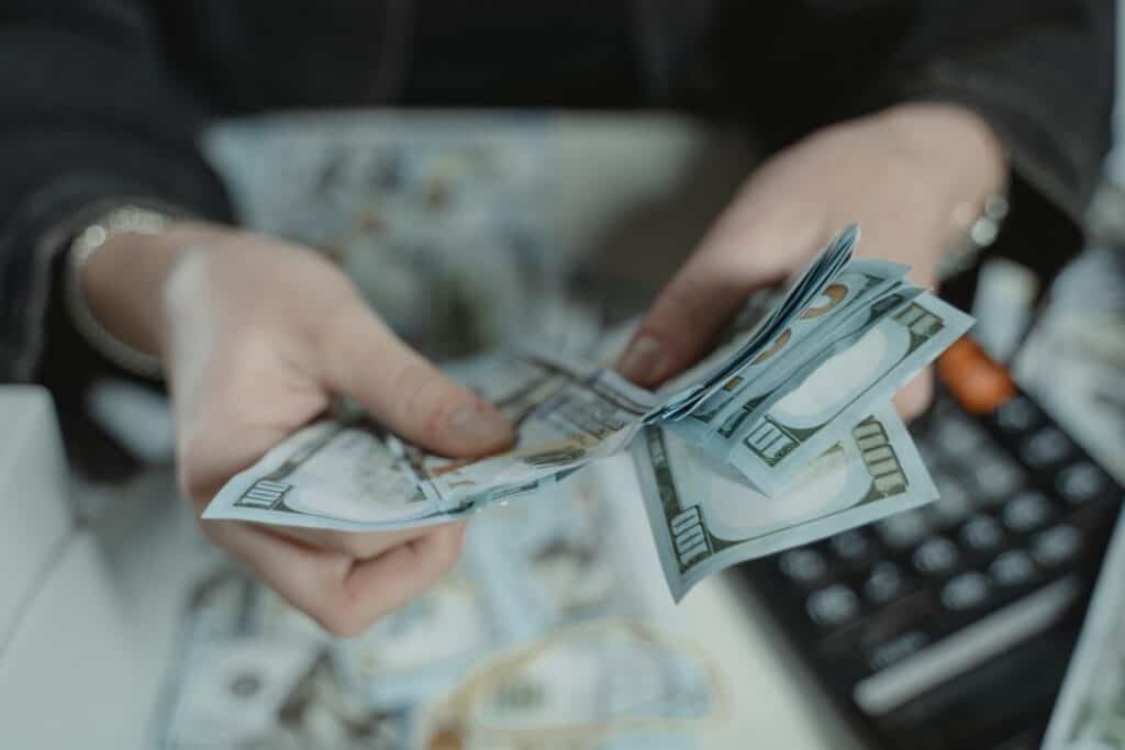 Close-up of hands counting a stack of US hundred-dollar bills, with more bills and a calculator on a surface.