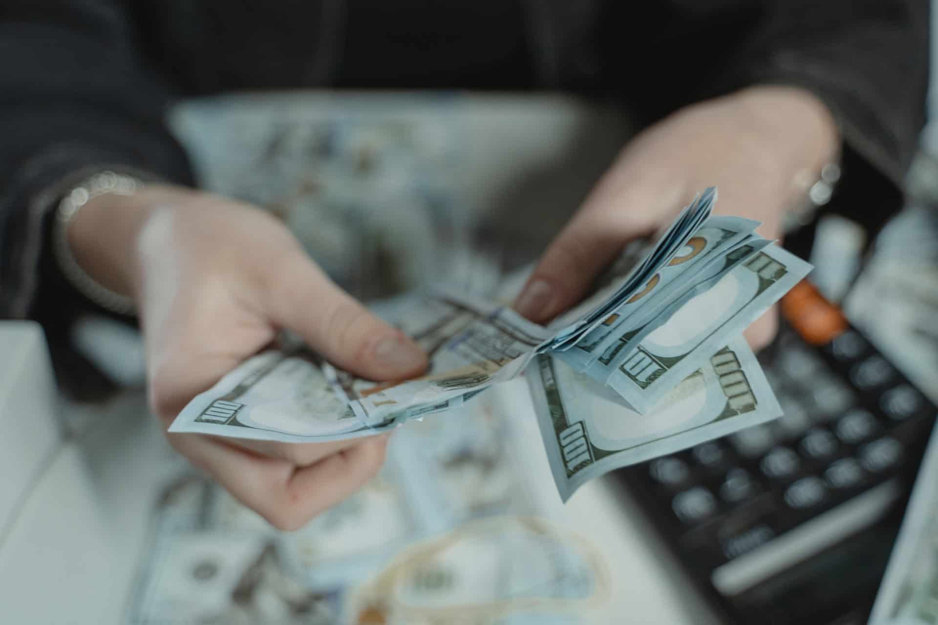 Close-up of hands counting a stack of US hundred-dollar bills, with more bills and a calculator on a surface.