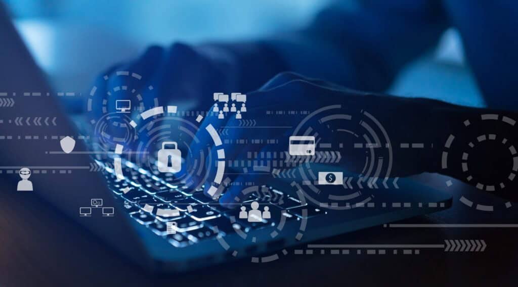 Hands typing on a laptop keyboard with overlaid digital icons representing cybersecurity, including a padlock, computer, credit card, dollar sign, and hacker symbol, all in a blue-toned, futuristic setting.