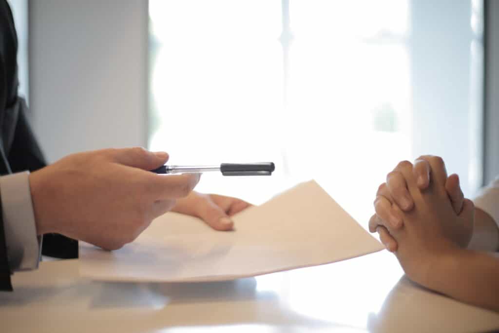 Close-up of two people at a table; one holds a pen over documents, the other has clasped hands.