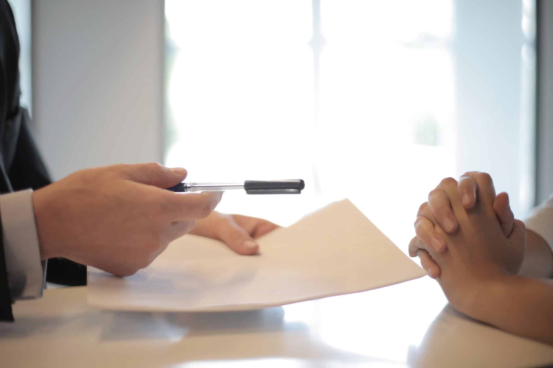 Close-up of two people at a table; one holds a pen over documents, the other has clasped hands.