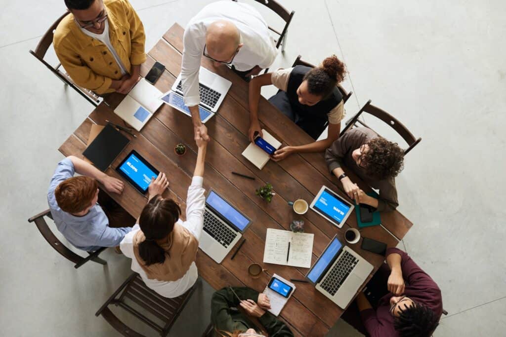 Overhead view of eight people around a wooden table, two shaking hands, surrounded by laptops, tablets, and notebooks.