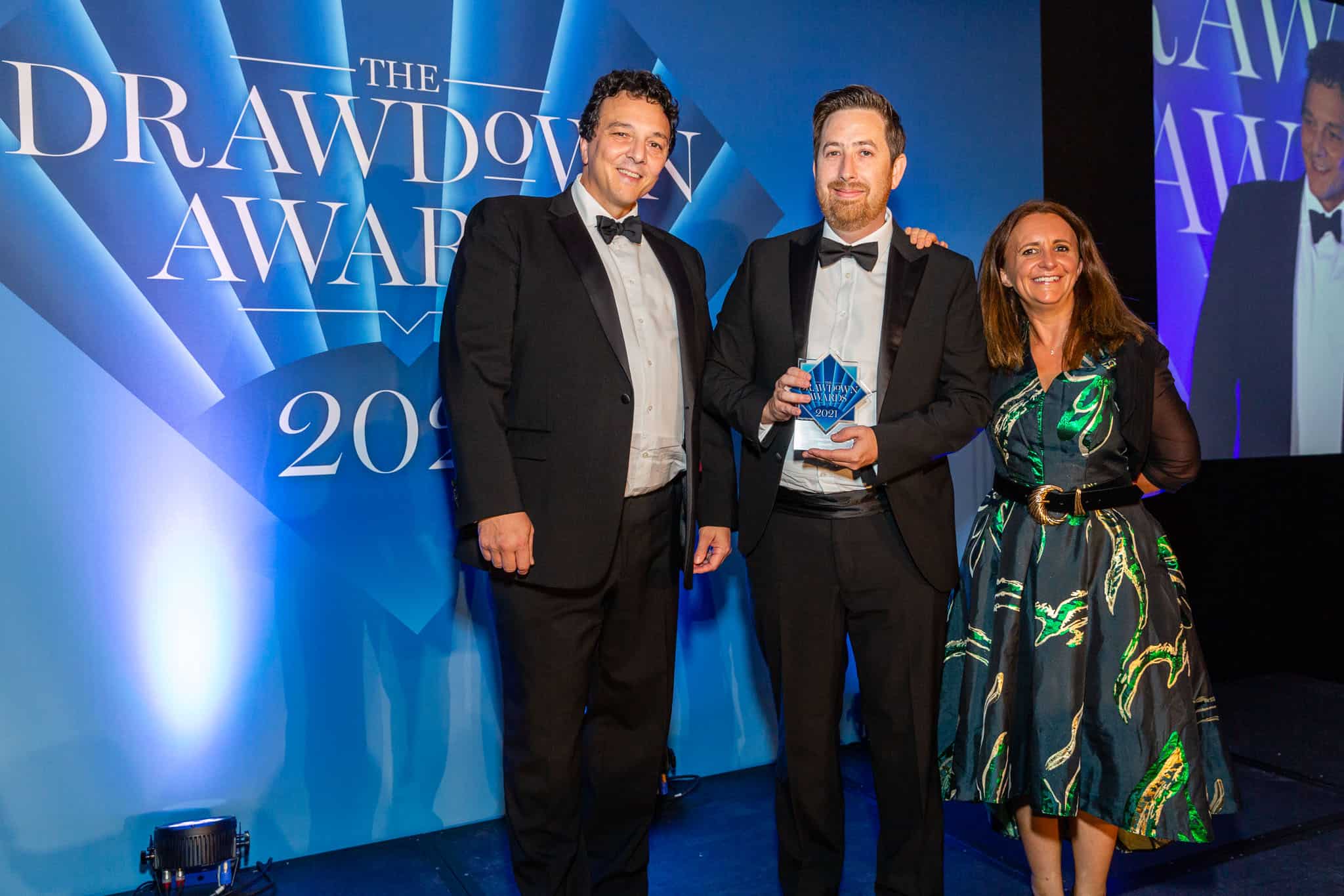 Two men in tuxedos and a woman in a patterned dress stand on stage, holding a star-shaped award at The Drawdown Awards 2021.
