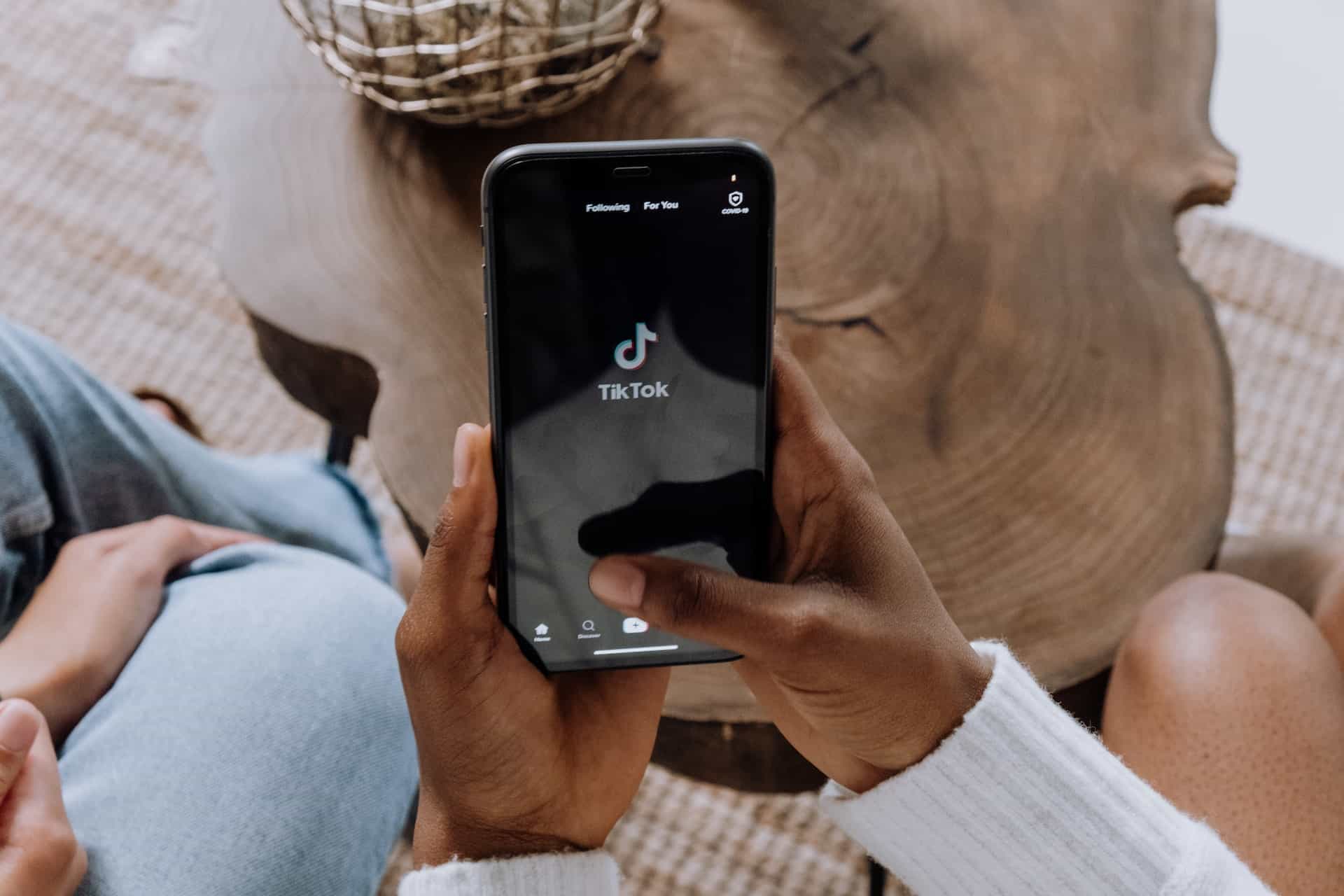 A person holds a black smartphone displaying the TikTok app logo on a dark screen. Hands, light blue jeans, a wooden table, and a woven rug are visible.