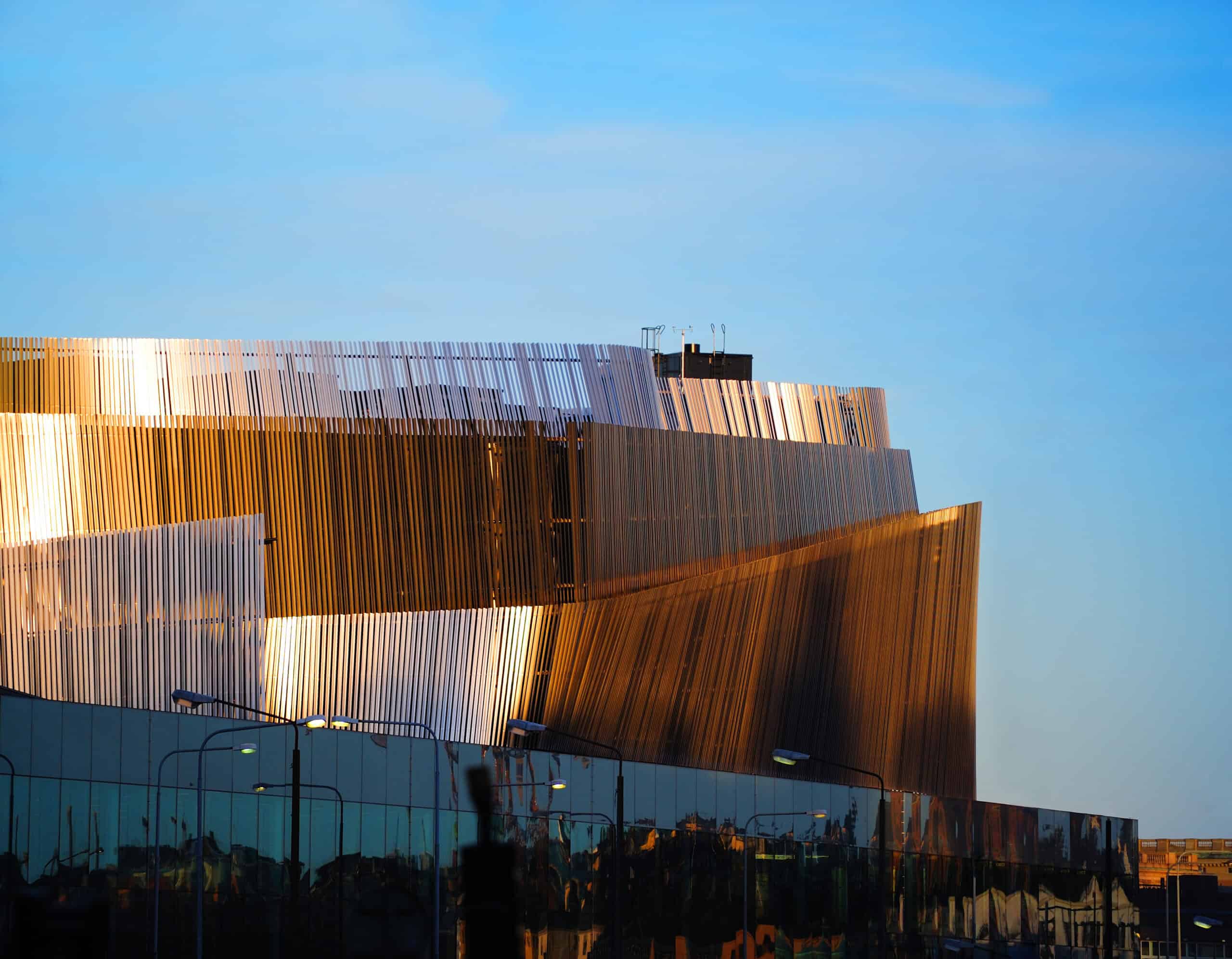 A modern building with a tiered, slatted facade. Sunlight illuminates parts of the slats in golden and orange, while other sections are in shadow. Below, reflective glass panels show distorted reflections. A clear blue sky is in the background.