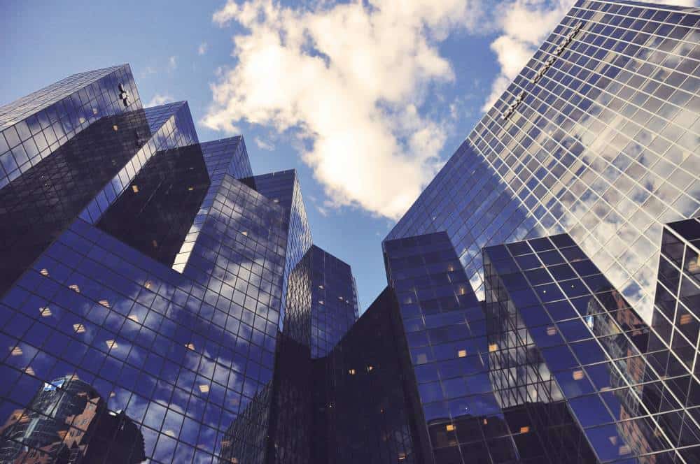 Low-angle view of multiple modern skyscrapers with dark blue reflective glass facades, reflecting a blue sky with white clouds.