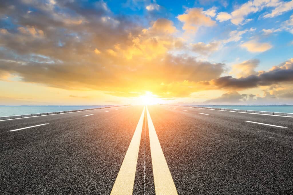 A long, straight asphalt road with yellow center lines extends towards a bright orange and yellow sunset over a body of water.