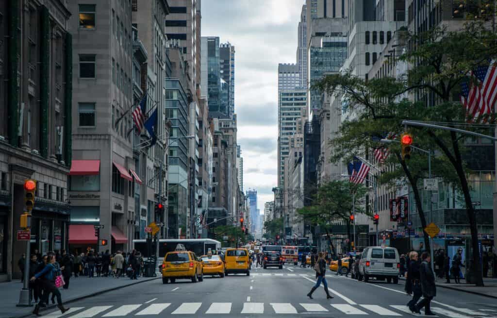 A wide shot of a busy city street with yellow taxis, pedestrians, and tall buildings under a cloudy sky. American flags are visible on some buildings.