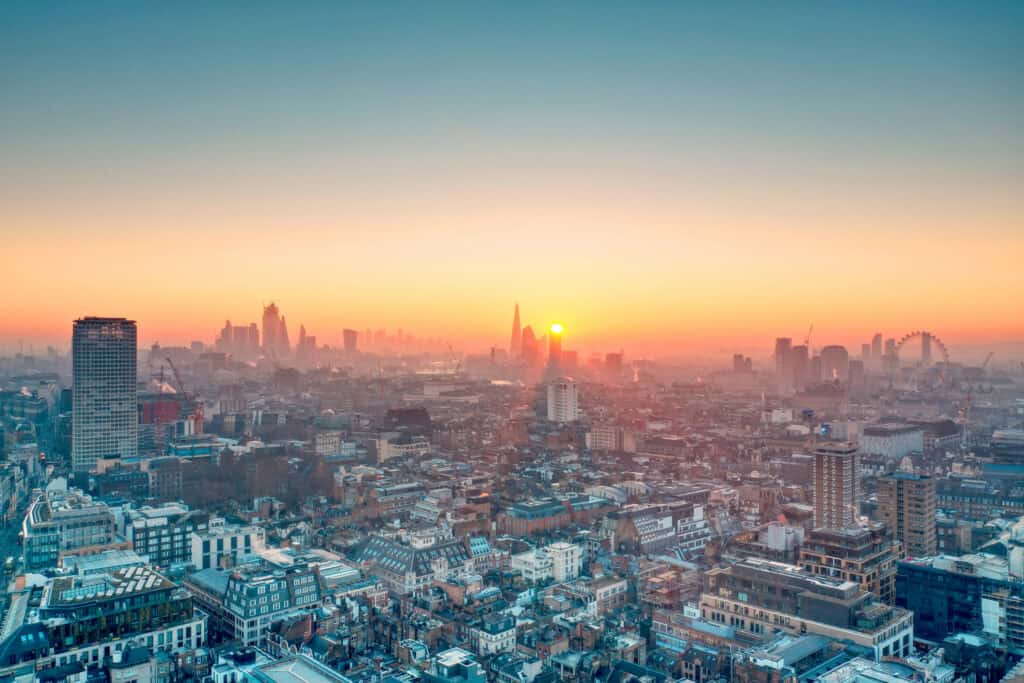 Panoramic aerial view of London at sunrise or sunset, showing a hazy cityscape with The Shard, other skyscrapers, and the London Eye under a gradient sky.