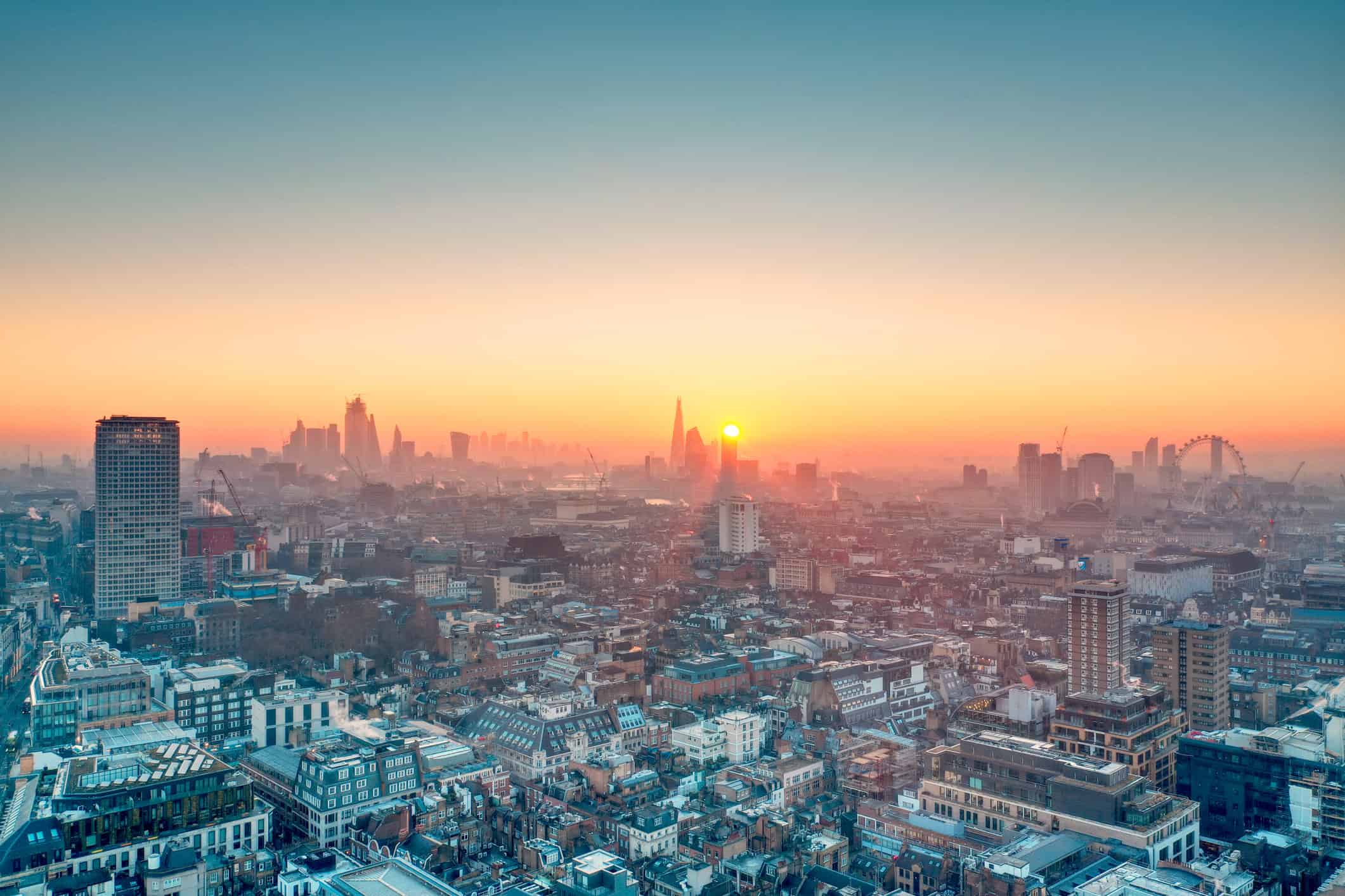 Panoramic aerial view of London at sunrise or sunset, showing a hazy cityscape with The Shard, other skyscrapers, and the London Eye under a gradient sky.