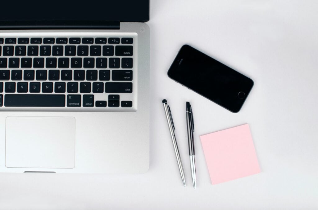 A top-down view of a silver laptop, black smartphone, two silver pens, and a pink sticky note on a white surface.