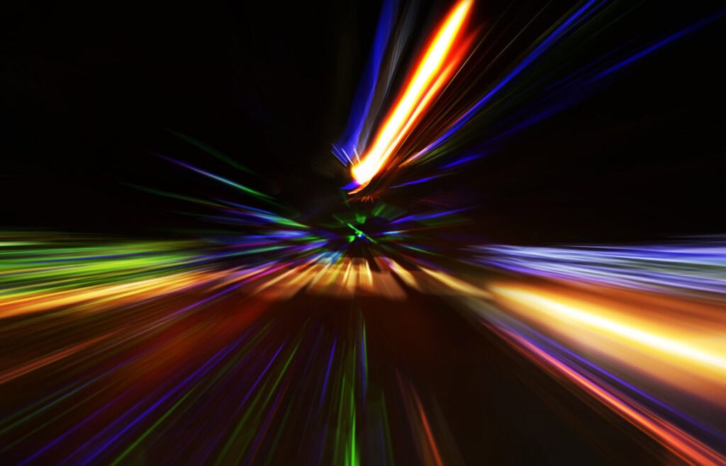 Abstract image of blurred, colorful light streaks in orange, yellow, red, green, blue, and purple, radiating from a central point on a black background.