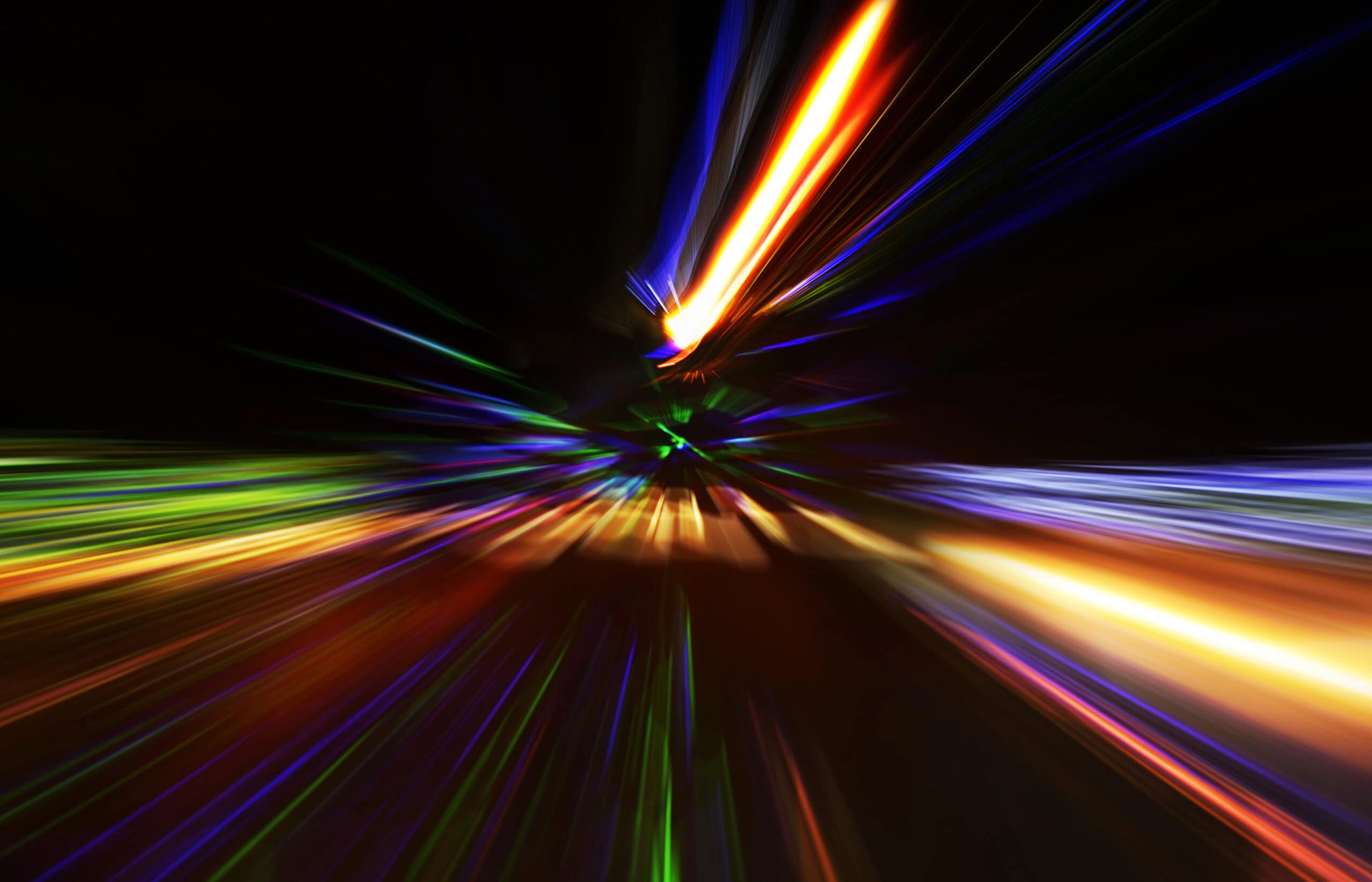 Abstract image of blurred, colorful light streaks in orange, yellow, red, green, blue, and purple, radiating from a central point on a black background.