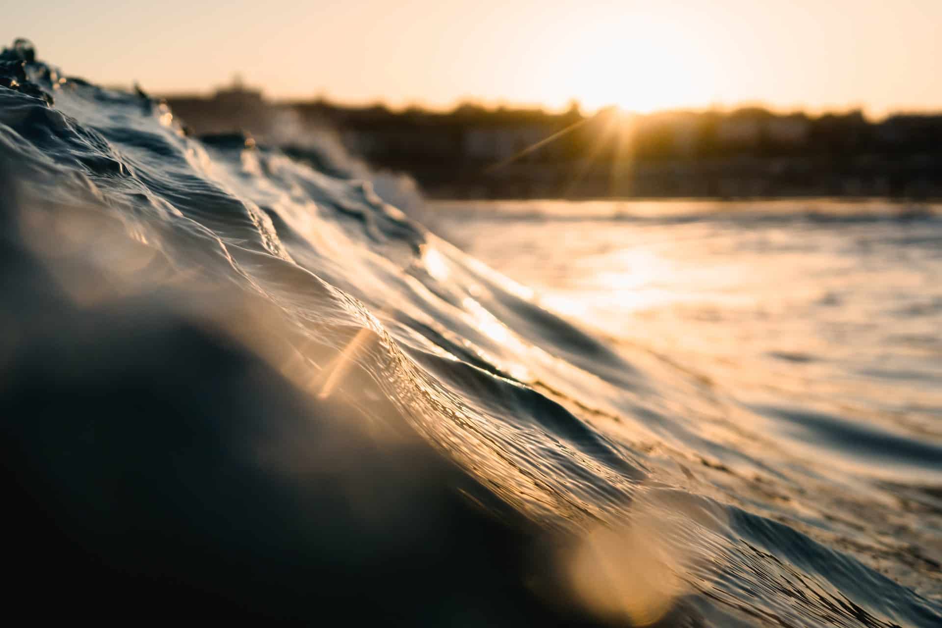Close-up of a textured ocean wave reflecting golden sunlight, with a sunburst and blurred distant land in the background.