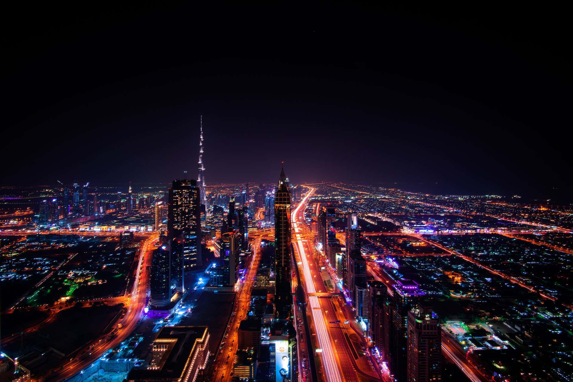 Aerial long-exposure shot of Dubai at night, showing the Burj Khalifa, illuminated skyscrapers, and streaks of red and white car lights on highways.