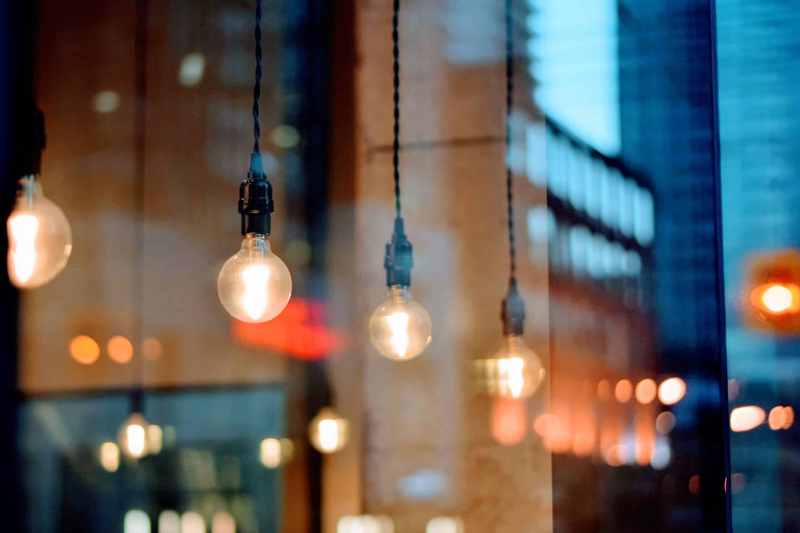 Four glowing Edison light bulbs hanging from wires, with blurry warm and cool reflections in the background.