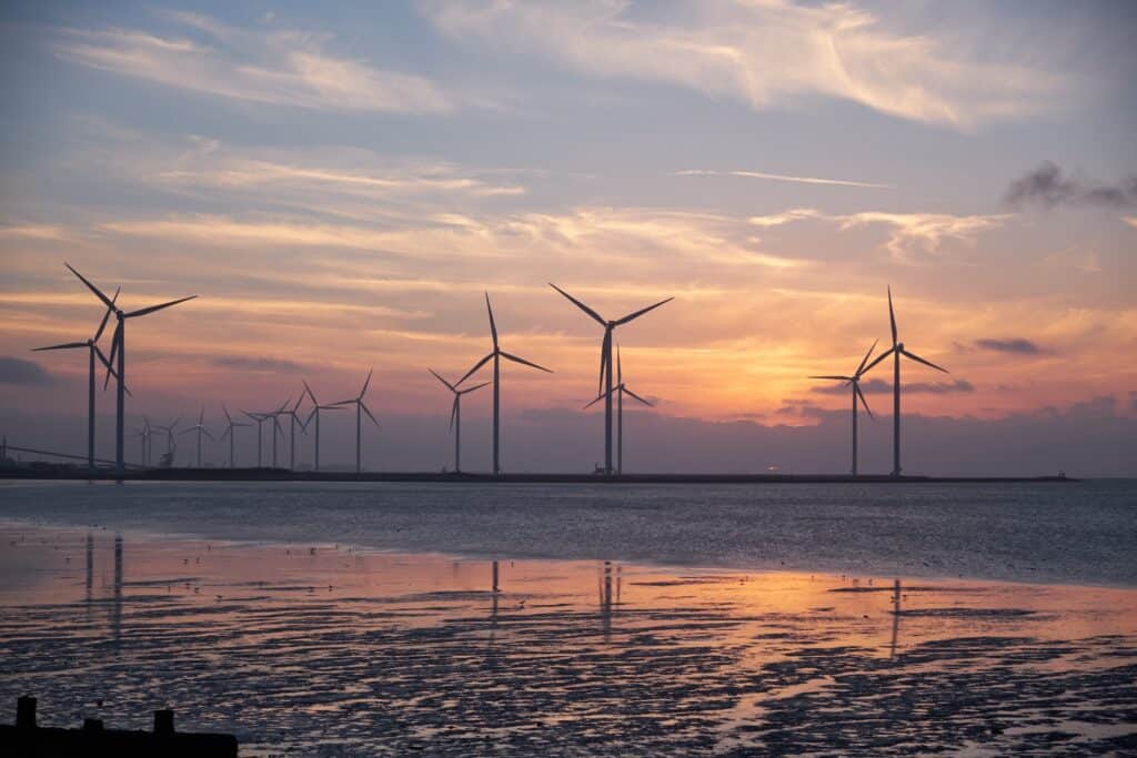 Silhouetted wind turbines stretching across the horizon at sunset, with a colorful sky reflected in the rippled water below.