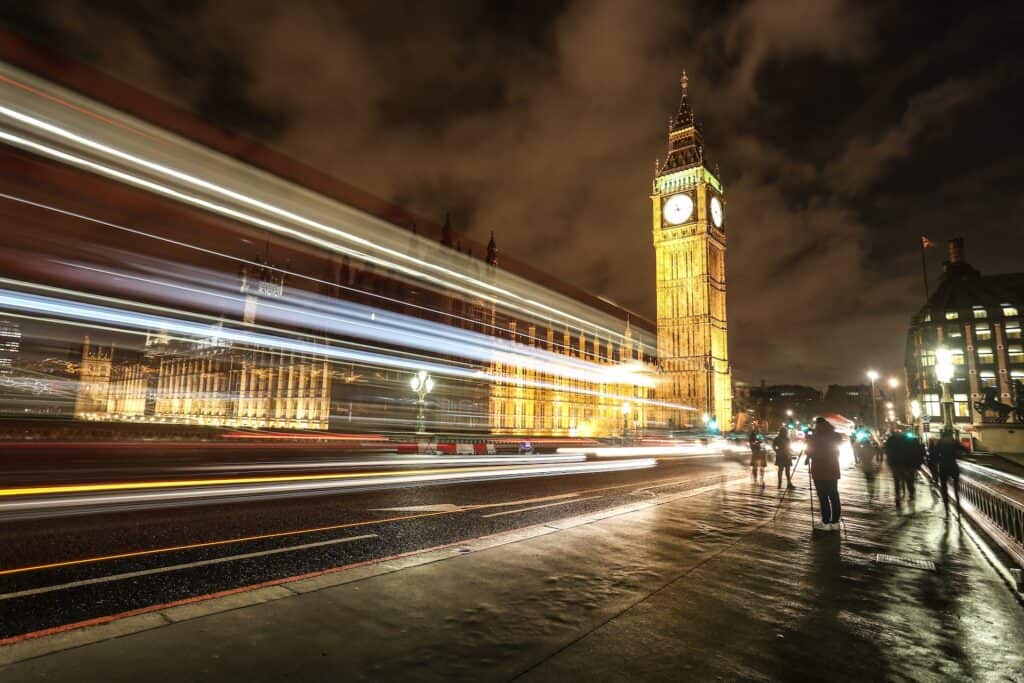 Long exposure night photo of Big Ben and Houses of Parliament, with bright light trails from vehicles and blurred figures on a wet sidewalk.