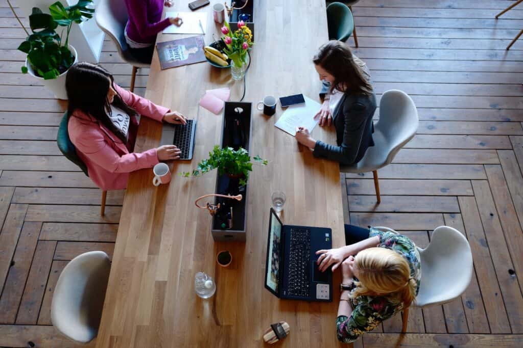 Overhead view of four people working at a long wooden table with laptops, papers, plants, and mugs.