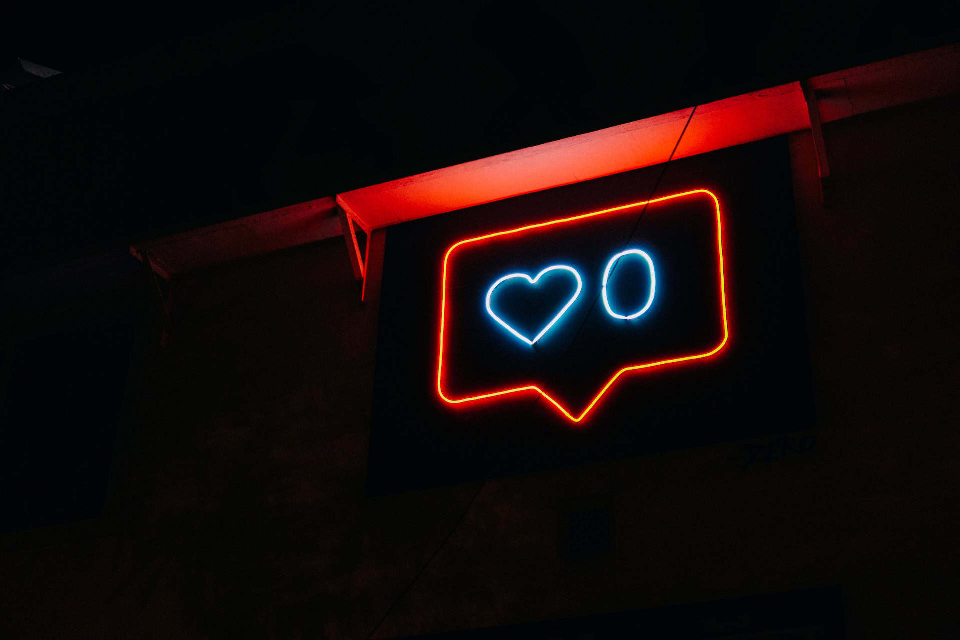 A neon sign in the shape of a social media 'like' icon, with an orange-red outline, a blue heart, and a blue circle, against a dark background.