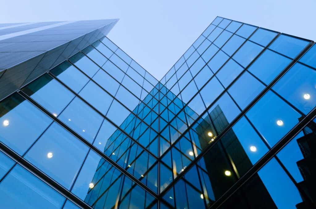 Low-angle view of a modern building with blue-tinted glass panels, forming a 'V' shape against a light blue sky. Reflections of interior lights are visible in the glass.
