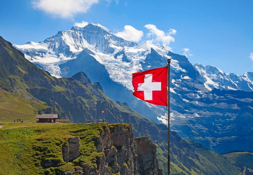 A large Swiss flag waving on a pole in front of majestic snow-capped mountains and green cliffs under a blue sky.