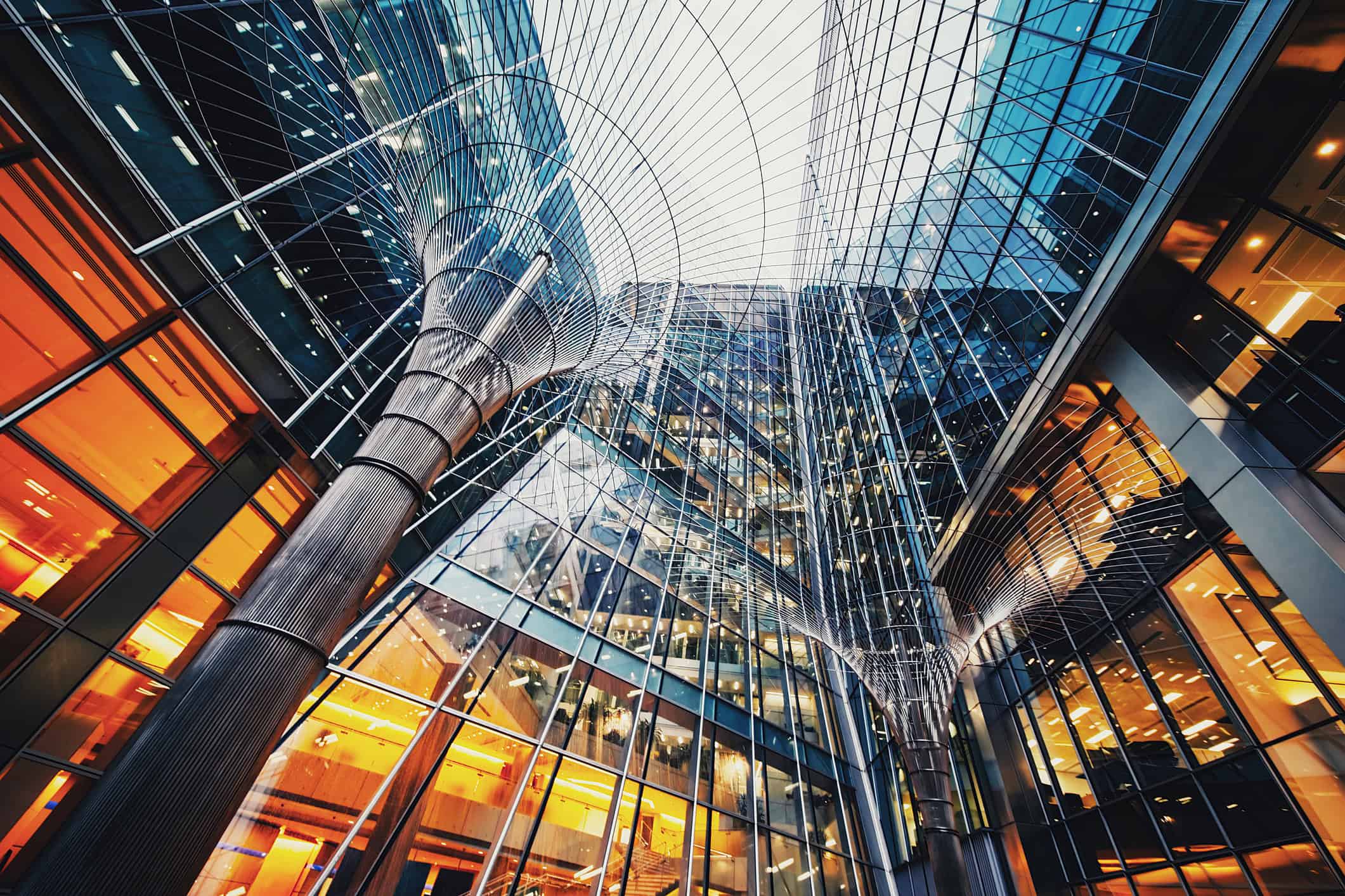 View looking up at a modern building interior with extensive glass walls reflecting orange and blue light, and a large ribbed column fanning into a complex grid structure overhead.