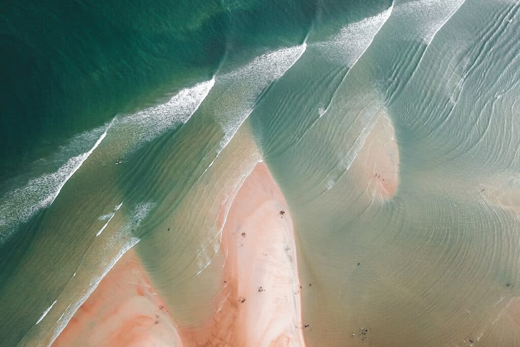 Aerial view of a beach with deep teal ocean, parallel lines of breaking white waves, and pinkish-orange sand, showing small figures and surfboards.