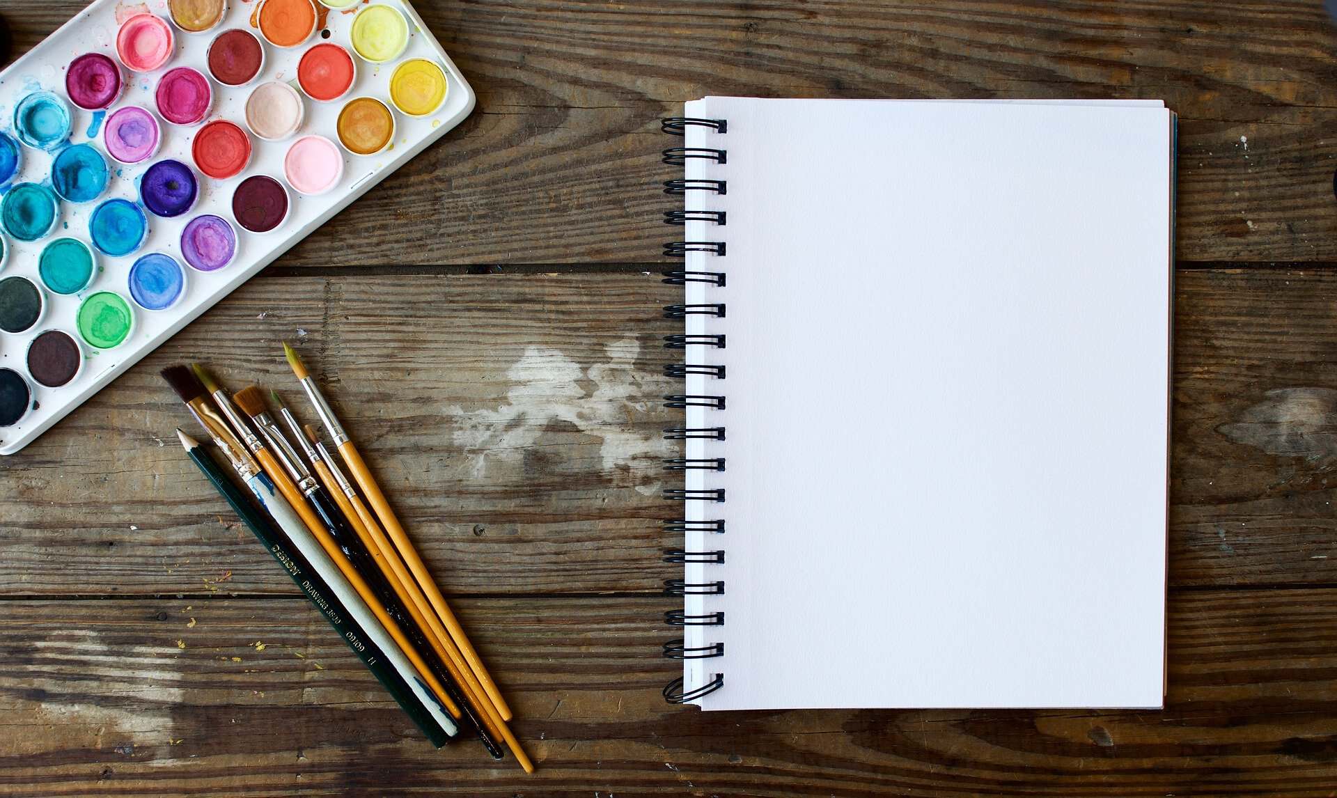 Overhead view of a watercolor palette, paintbrushes, and a blank spiral-bound sketchbook on a wooden surface.