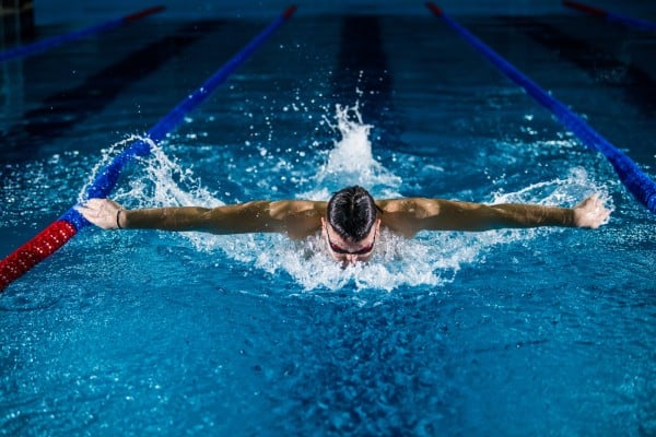 A male swimmer performing the butterfly stroke in a blue swimming pool, creating splashes with his arms extended.