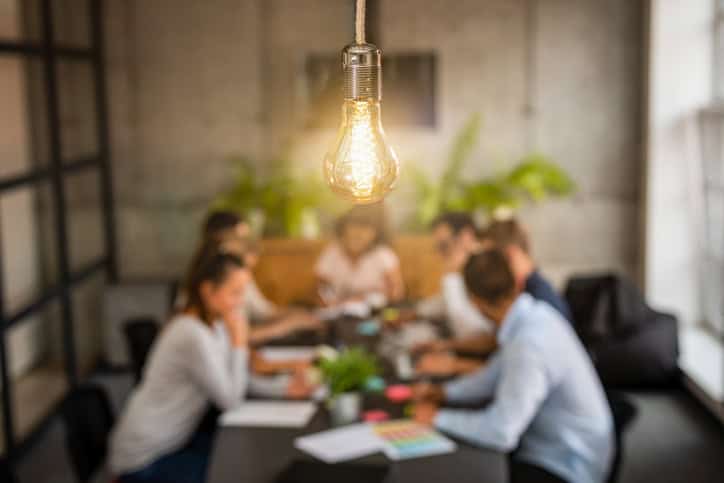 A glowing vintage light bulb hangs in the foreground, with a blurred group of people in a meeting around a table in the background.