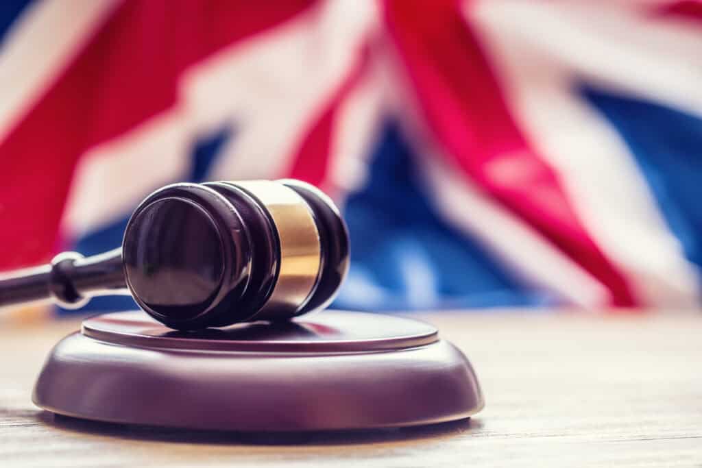 A dark wooden gavel with a brass band resting on a wooden surface, with a blurred Union Jack flag in the background.
