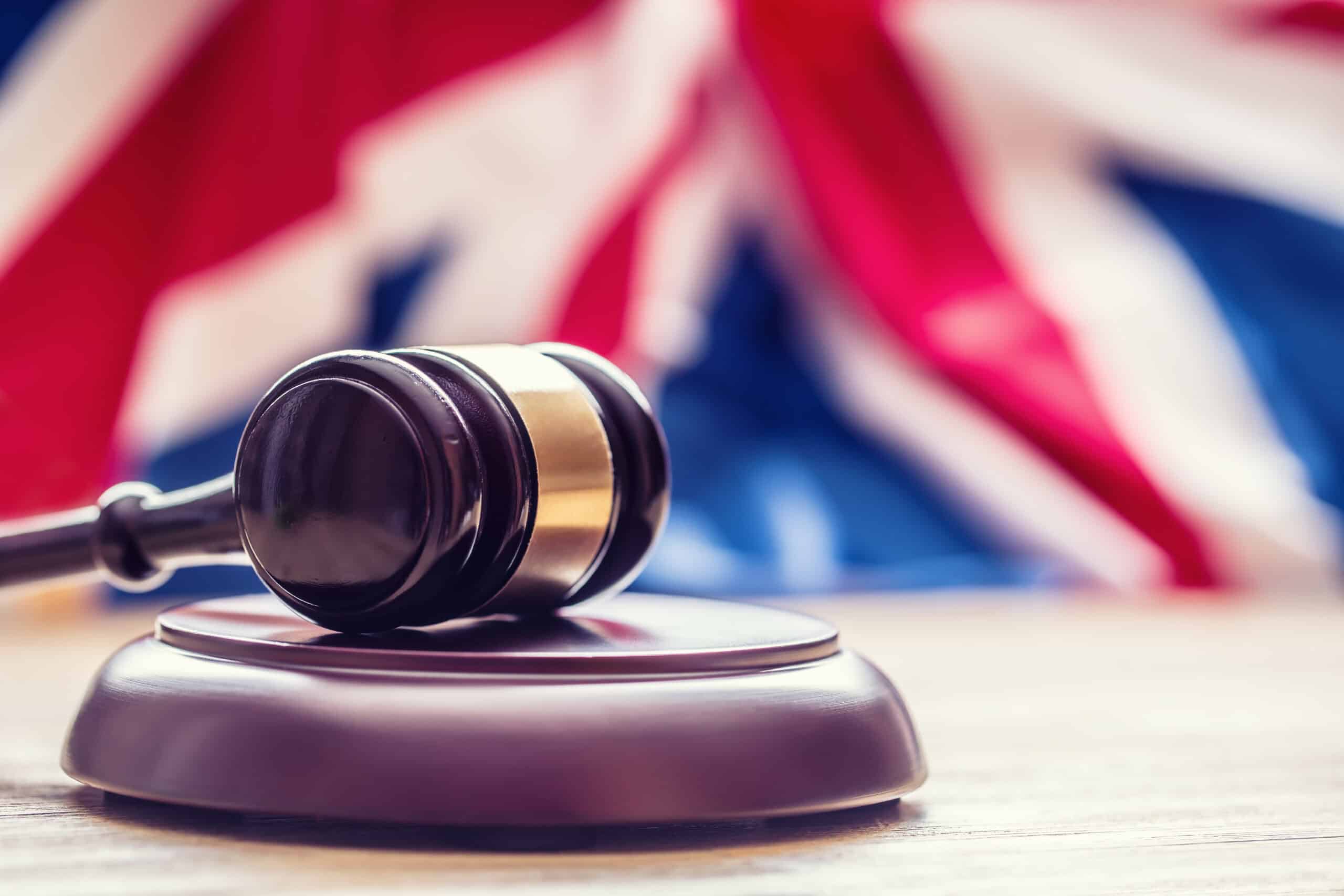 A dark wooden gavel with a brass band resting on a wooden surface, with a blurred Union Jack flag in the background.