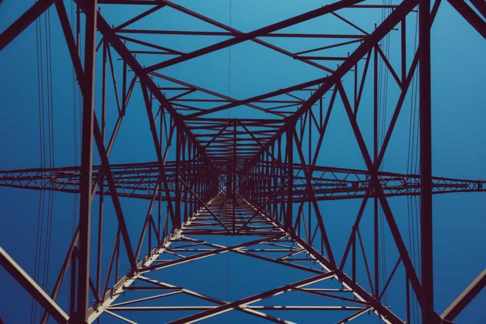 Low-angle view looking up into the metal framework of a power line tower against a clear blue sky.