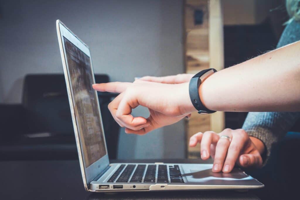 Close-up of hands interacting with a silver laptop. One hand points at the screen, while the other rests near the trackpad. A black fitness tracker is on one wrist, and a silver ring is on a finger.