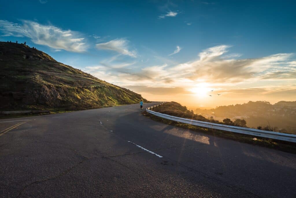 A person runs on a winding asphalt road around a green hill at sunset, with a bright golden sky, distant town, and three birds in flight.