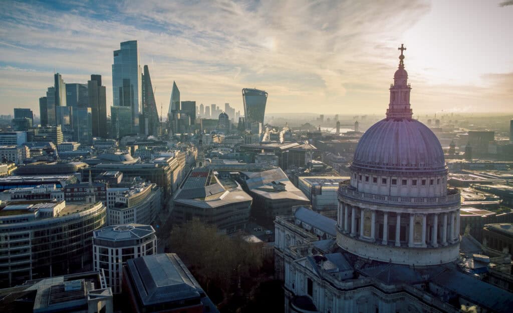 Aerial view of London's cityscape, featuring St. Paul's Cathedral dome in the foreground and a cluster of modern skyscrapers under a partly cloudy sky.