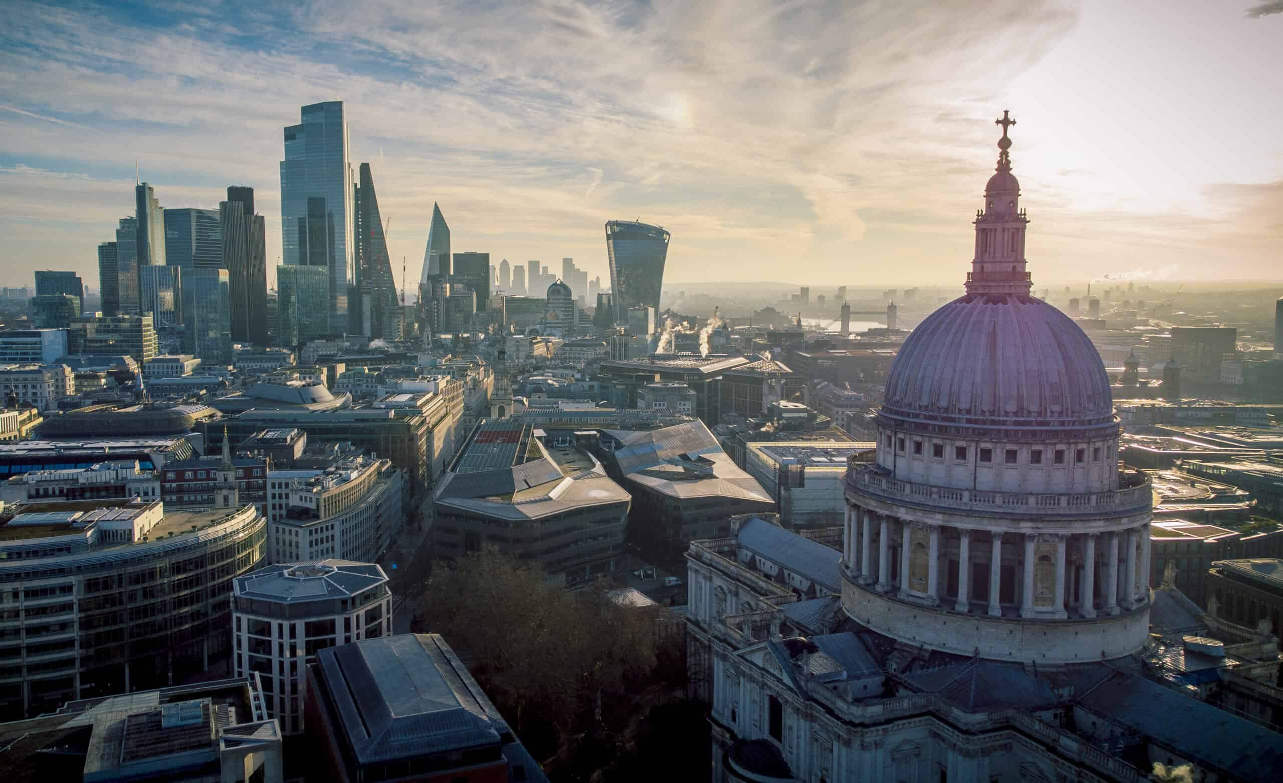 Aerial view of London's cityscape, featuring St. Paul's Cathedral dome in the foreground and a cluster of modern skyscrapers under a partly cloudy sky.