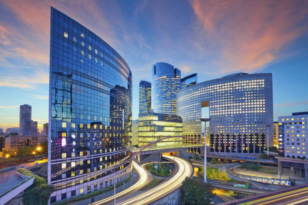 A long-exposure shot of a modern city at dusk, featuring curved glass skyscrapers, a complex highway interchange with light trails, and a sky with blue, pink, and orange clouds.