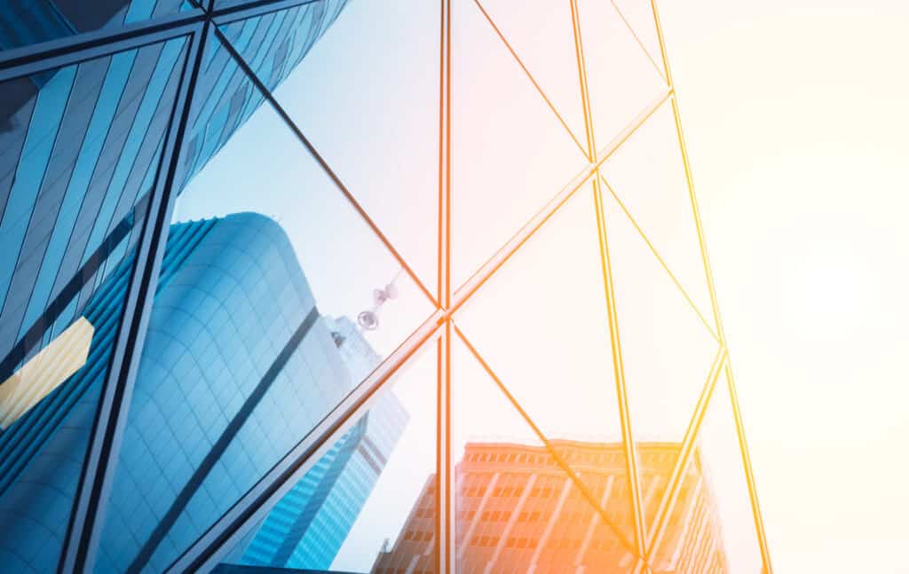 Close-up of a modern glass building facade reflecting other buildings and a bright sky, with a cool blue tint on the left and a warm orange-yellow glow on the right.