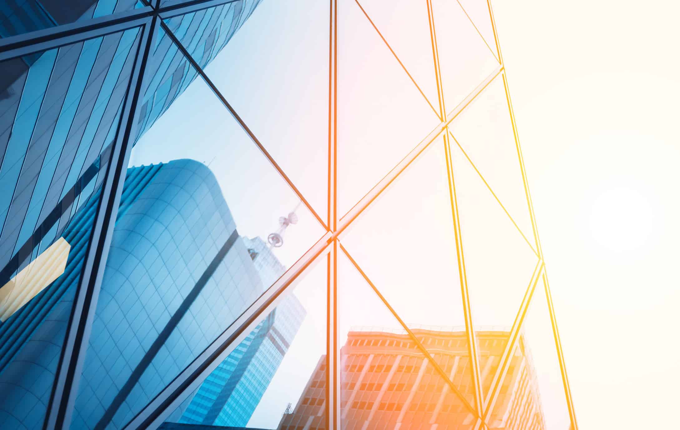 Close-up of a modern glass building facade reflecting other buildings and a bright sky, with a cool blue tint on the left and a warm orange-yellow glow on the right.