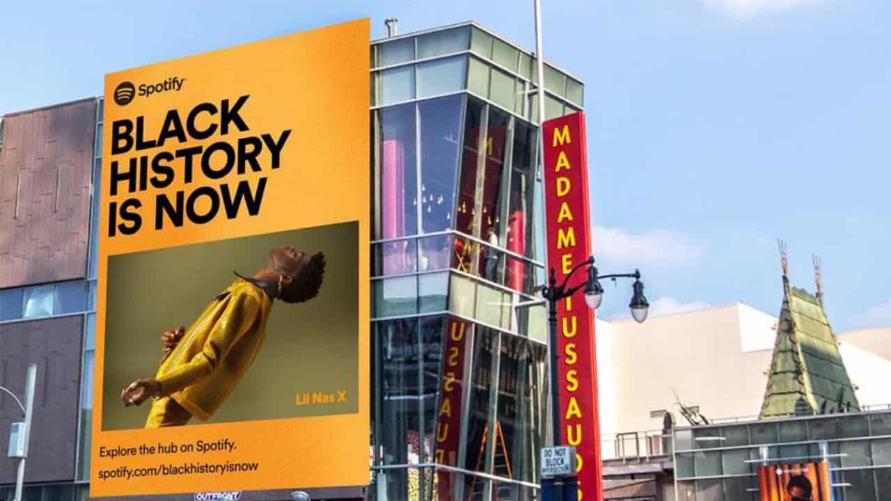 A large orange billboard for Spotify's 'Black History Is Now' campaign featuring Lil Nas X in a yellow jacket, next to a Madame Tussauds building.