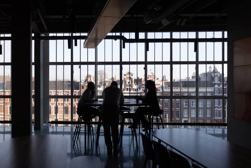 Three people silhouetted at a high table in front of large windows overlooking a city skyline with traditional buildings.