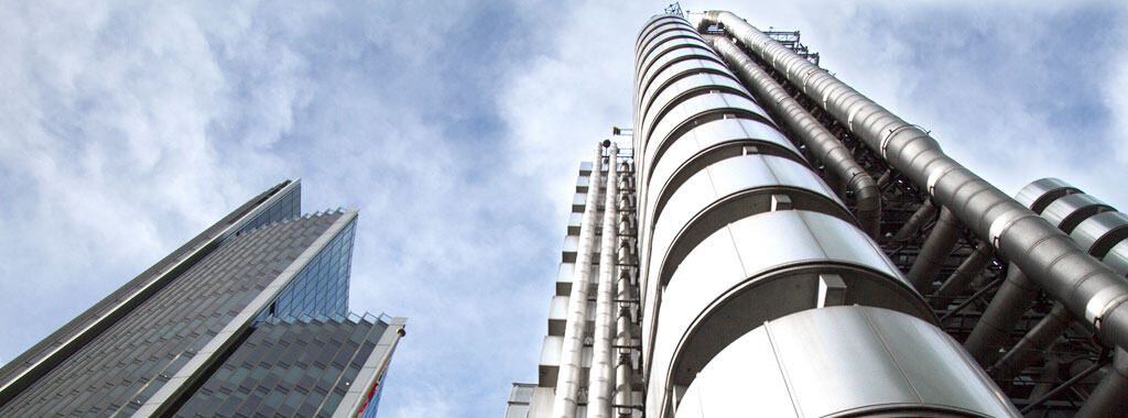Two modern skyscrapers against a cloudy sky. The building on the right is the Lloyd's building with external pipes, and the one on the left is a glass and steel tower.